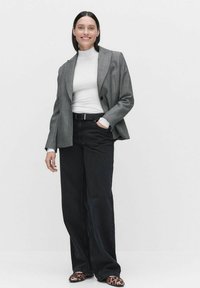 Grijze, op maat gemaakte blazer over een witte coltrui, gecombineerd met zwarte wijde jeans en leoprint instappers.