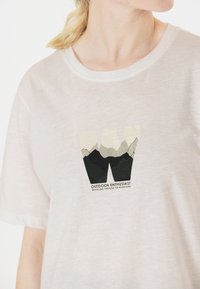 Vit t-shirt med kort ärm som har ett motiv av berg och texten "UTOMHUSENTUSIAST" i svarta, gröna och beige färger.