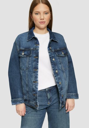 Donkerblauwe denim spijkerjas met zilverkleurige knopen aan de voorkant, twee borstzakken en een klassieke kraag. Ggedragen over een wit topje.