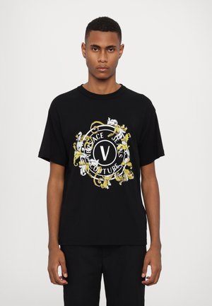 Črna bombažna majica z velikim, pisanem grafičnim motivom z zlatimi in belimi cvetličnimi vzorci ter napisom "VERSACE JEANS COUTURE" v krožni zasnovi.