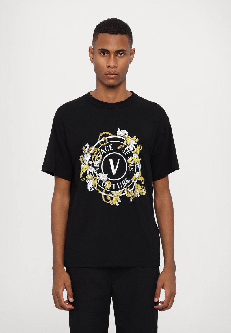 Juodas medvilninis marškinėlis su dideliu, spalvingu grafiniu motyvu, puoštas auksiniais ir baltais gėlių raštais, su tekstu "VERSACE JEANS COUTURE" apvalioje kompozicijoje.