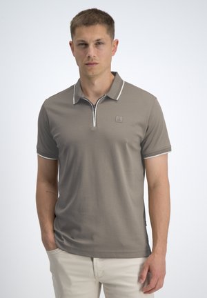 Giovane uomo in piedi con le mani parzialmente nelle tasche, indossa una polo maniche corte color taupe con bordo bianco e pantaloni chiari.