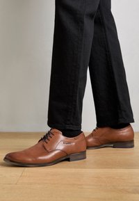 Chaussures habillées en cuir marron à finition lisse, à bout arrondi, avec des lacets noirs. La semelle est d'une teinte foncée contrastante, assortie à un pantalon noir.