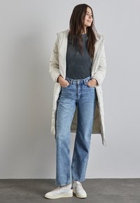 Gevoerde lange jas in off-white, geribbelde donkergrijze top, lichtblauwe rechte jeans en witte sneakers met gemarmerde accenten.