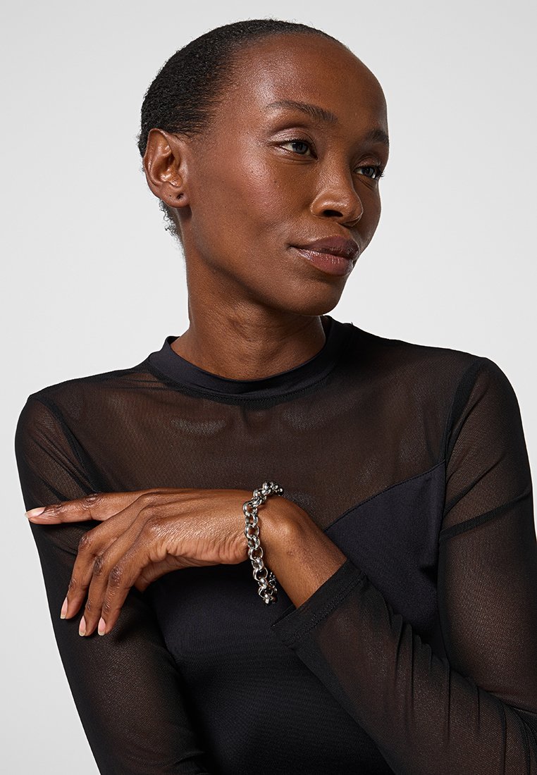 Top lungo nero trasparente con un design geometrico; bracciale in argento con maglie rotonde indossato al polso. Pelle liscia e trucco naturale.