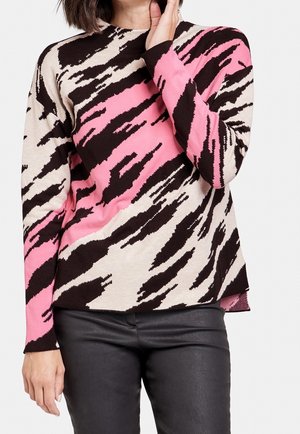 Kobieta ubrana w sweter z długim rękawem w różowo-czarno-beżowe abstrakcyjne paski tygrysa, zestawiony z czarnymi skórzanymi spodniami, ręka przy twarzy.