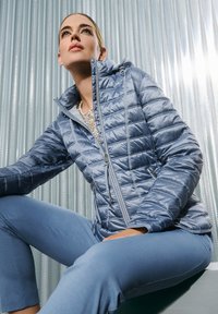 Himmelblaues, gepolstertes Jacket mit glänzender Oberfläche, ausgestattet mit Kapuze, Reißverschluss vorne und zwei Taschen. Tragen mit passenden blauen Hosen.