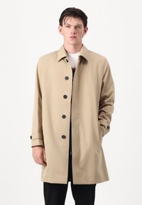 Beige, lightweight coat with a straight cut, featuring a collar, black buttons, and cuff detail. Worn over a white graphic tee.