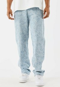 Jeans de color azul claro con patrones de paisley blancos, combinados con zapatillas blancas y una camisa blanca, mostrados desde la cintura hasta los pies.