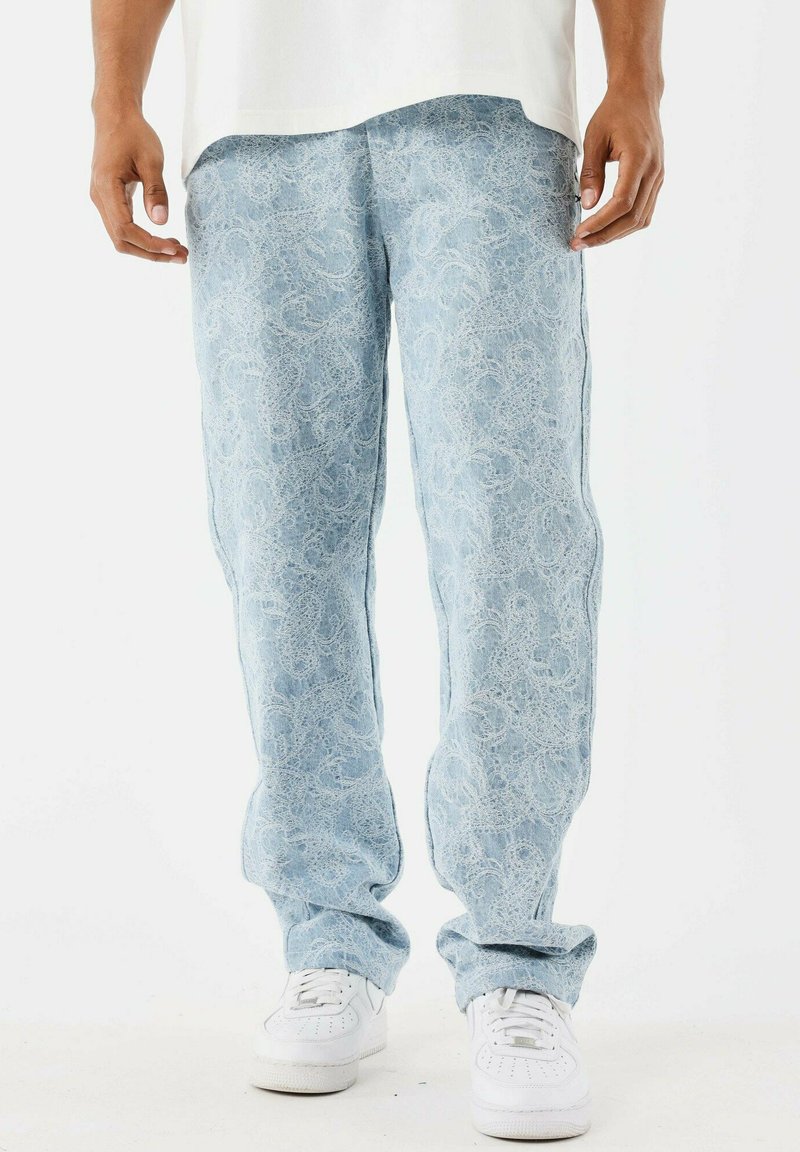 Jeans de color azul claro con patrones de paisley blancos, combinados con zapatillas blancas y una camisa blanca, mostrados desde la cintura hasta los pies.