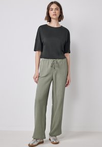 Dark green short-sleeve top paired with light green, textured wide-leg pants. Model wears silver sneakers, standing against a neutral background.