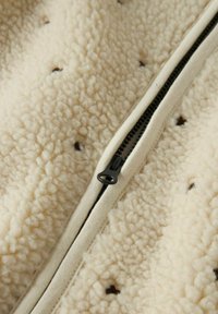 Veste en polaire crème, avec une surface texturée, une fermeture éclair noire et de petits points bruns comme détail sur le tissu.