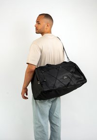 Black duffel bag with a textured finish, secure side handles, and elastic bungee accents. Rear view with adjustable shoulder strap.