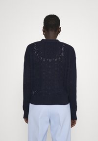 Personne aux cheveux courts portant des boucles d'oreilles en or, un pull en maille bleu marine avec un motif en dentelle et un pantalon bleu clair, vue de dos.