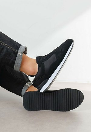 Personne portant des baskets noires et blanches avec des semelles crantées, jambes croisées, jean noir retroussé contre un fond clair uni.