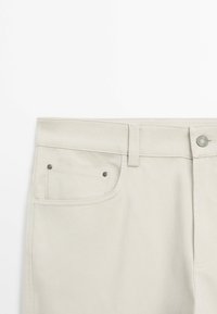 Vue de face d'un pantalon beige clair montrant la taille, la passants de ceinture, le bouton métallique et la poche avant avec rivets.