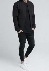 Giubbotto bomber nero con zip frontale, abbinato a jeans skinny neri strappati e sneakers bianche alte. Texture liscia e design aderente.