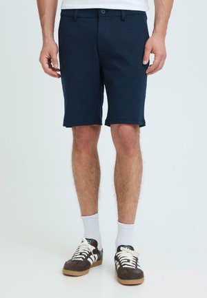 Mann iført marineblå shorts til knærne, hvite sokker og svarte og hvite joggesko, stående mot en ensfarget lys bakgrunn.