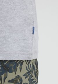 Camiseta de algodón gris con una textura lisa, que presenta una pequeña etiqueta azul en el lateral. Combinada con pantalones cortos verdes, que muestran un estampado floral.