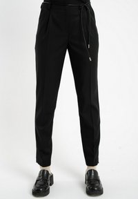 Czarne zwężające się spodnie z cienkim czarnym paskiem, noszone z beżowymi skarpetkami i czarnymi masywnymi półbutami na białym tle.