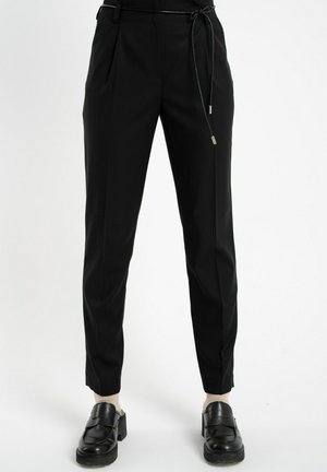 Czarne zwężające się spodnie z cienkim czarnym paskiem, noszone z beżowymi skarpetkami i czarnymi masywnymi półbutami na białym tle.