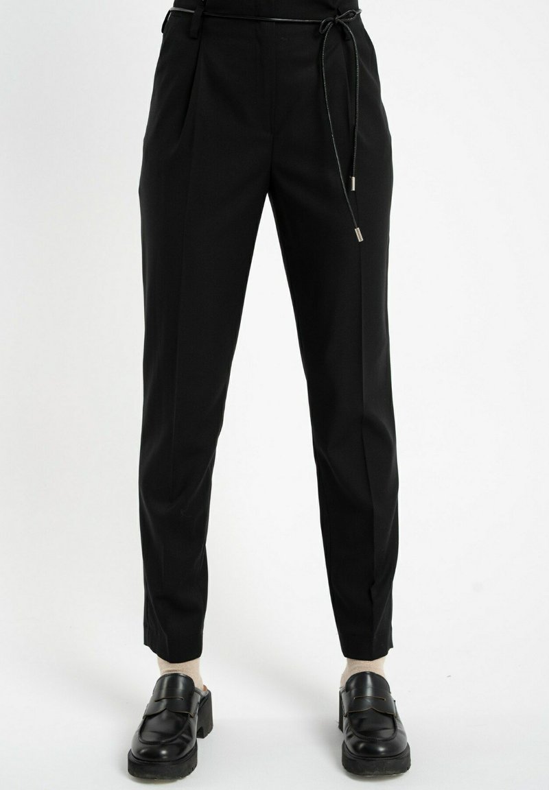 Czarne zwężające się spodnie z cienkim czarnym paskiem, noszone z beżowymi skarpetkami i czarnymi masywnymi półbutami na białym tle.