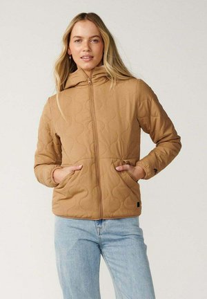 Junge Frau trägt eine beige gesteppte Jacke mit Vordertaschen und hellblaue Jeans, steht vor einem neutralen Hintergrund.