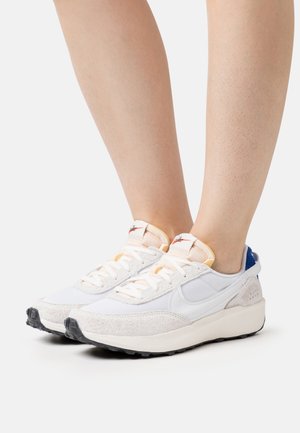 White athletic sneakers with beige suede accents, mesh upper, black rubber outsole, and orange detailing on the tongue and heel.