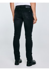 Férfi, aki fekete szűk fazonú farmert és fekete bőrcsizmát visel, hátulról nézve világos, egyszerű háttér előtt áll.