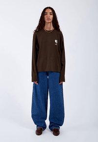 Brun langærmet t-shirt med hvidt hjerteprint, kombineret med vide blå denimjeans, vist på en model. Glat stoftekstur.