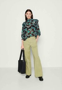 Transparante zwarte blouse met teal en witte bloemenpatroon, met gerimpelde mouwen; gecombineerd met olijfgroene flared broek; model houdt een zwarte tas vast.