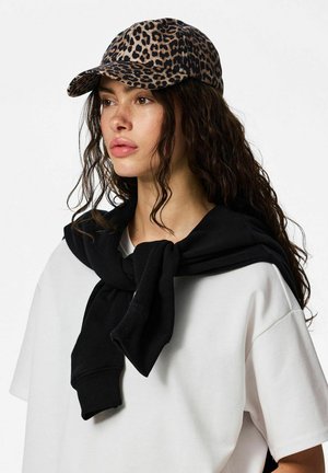 Joven con cabello largo y rizado que lleva una gorra de béisbol con estampado de leopardo, camiseta blanca y un suéter negro atado alrededor de los hombros.