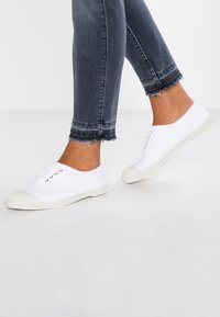 Sneakers bianchi in tela con punte e occhielli in gomma, caratterizzati da un design semplice. Abbinati a jeans in denim scuro con risvolti.