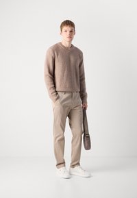 Hellbrauner strukturierter Pullover mit V-Ausschnitt, kombiniert mit beigen Hosen und weißen Sneakers. Eine graue Umhängetasche wird in einer Hand gehalten.
