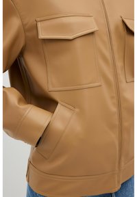 Tan syntetisk læderjakke med to frontlommer, høj krave og lynlåslukning. Struktureret overflade med synlige syninger.