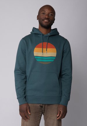 Teal hoodie met een voorzak en een trekkoord. Kenmerkt zich door een cirkelvormige afbeelding met oranje, geel, beige, turquoise en bruine strepen.