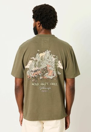 Mann mit kurzen lockigen Haaren trägt ein olivgrünes T-Shirt mit einem Design von Berg, Bäumen, Van und "Wild. Fast. Free. Volkswagen" auf dem Rücken.
