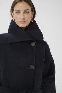 Tengerészkék gyapjú kabát magas gallérral, nagy gombokkal és öves derékkal. Puha textúra és szabott fazon, letisztult, minimál dizájnnal.