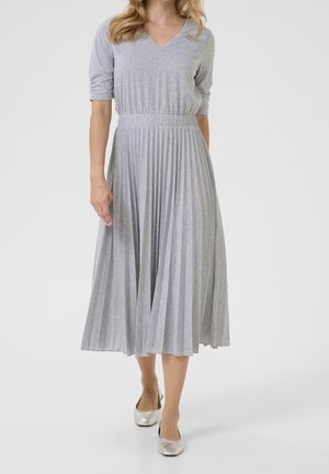 Femme portant une jupe midi plissée gris clair et un haut assorti à col en V avec manches 3/4, associée à des chaussures plates argentées, debout sur un fond uni.