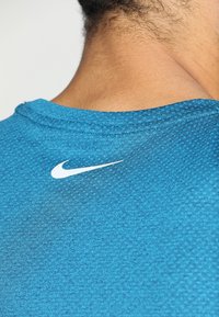 Turkoosi Nike-urheilupaita, jossa on teksturoitu kangas. Kaalta löytyy valkoinen logo, pyöreä kaulus ja sileä reuna.