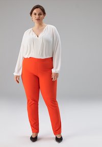 Weißes Langarmshirt mit Kordelzug am Ausschnitt, kombiniert mit enganliegenden roten Hosen; das Modell trägt schwarze spitze Ballerinas. Glatter, leichter Stoff.