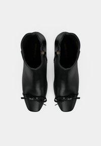 Botas de tornozelo em couro preto com uma textura suave, bico fino e detalhe em laço de cadarço. Apresentam um fecho de zíper dourado na lateral.