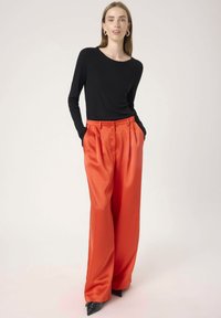 Un haut noir à manches longues associé à un pantalon en satin orange vif à jambes larges avec des plis, et des talons noirs pointus. Accessoires minimalistes visibles.