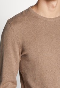 Maglione marrone a maglia con scollatura a girocollo, caratterizzato da una texture liscia e maniche aderenti, parzialmente visibile dalla spalla al collo.