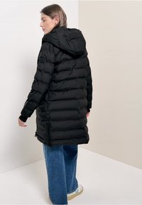 Sort puffjakke med hætte, med en quiltet design, elastiske manchetter og sidelommer. Sammen med bredbenede blå jeans og let fodtøj.