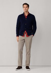 Cárdigan ribeteado azul marino con botones dorados, llevado sobre una camisa roja. Pantalones de cuadros en beige claro, zapatillas marrones con suelas blancas.