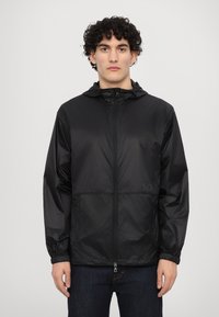Veste légère noire avec capuche, fermeture éclair à l'avant, poignets élastiques et détail de logo. Texture lisse, conçue pour un usage quotidien.