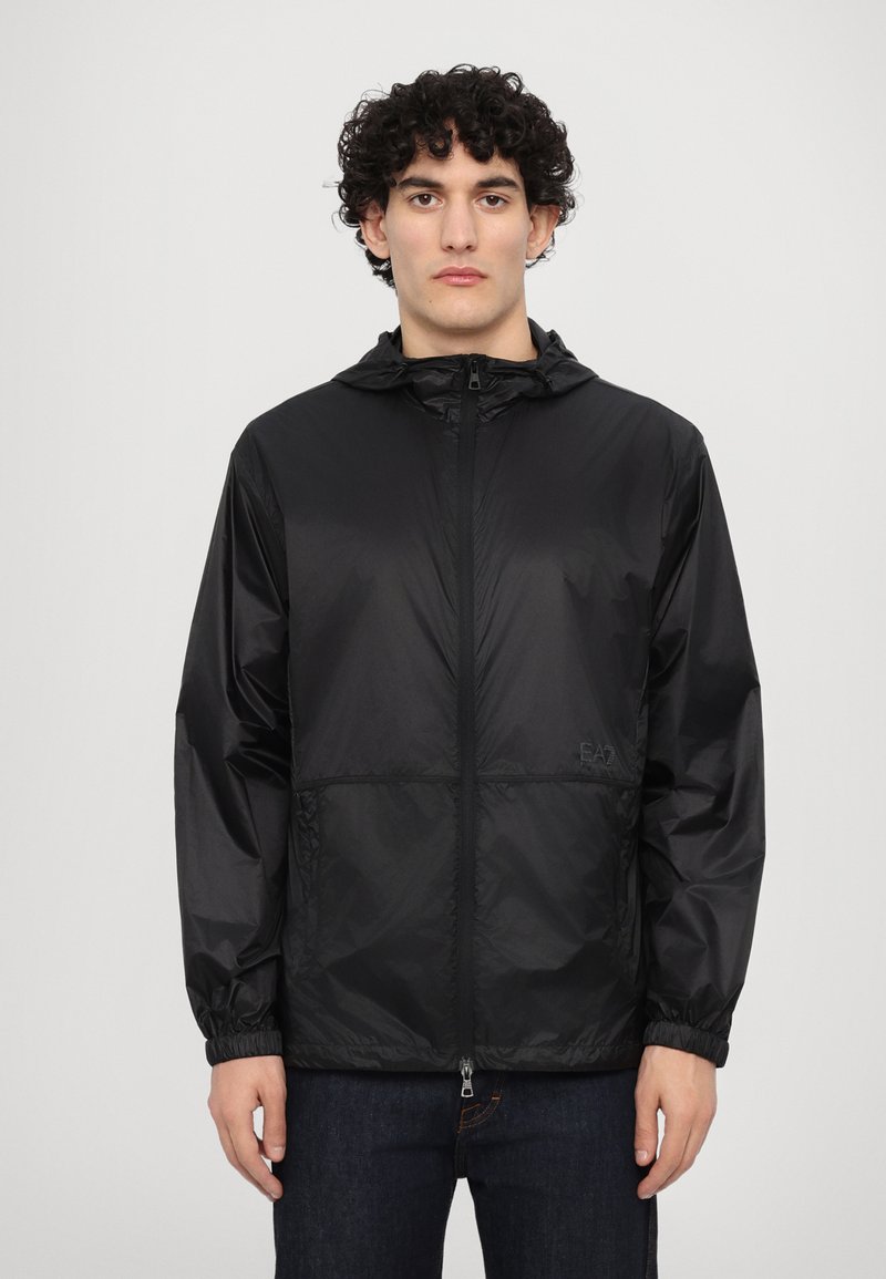 Veste légère noire avec capuche, fermeture éclair à l'avant, poignets élastiques et détail de logo. Texture lisse, conçue pour un usage quotidien.