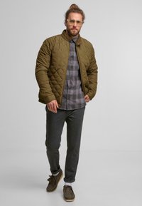 Vihreä tilkkupaita, jossa on pystykaulus, päällä harmaa ruutupaita, yhdistettynä tummanharmaisiin housuihin ja ruskeisiin jalkineisiin.