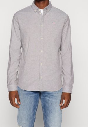 Homme portant une chemise à boutons gris clair avec un petit logo sur la poitrine et un jean bleu délavé, sur un fond uni.